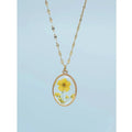 A gold-plated necklace with a clear resin-encased dried yellow flower pendant, presented against a light blue background.
