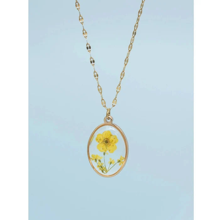 A gold-plated necklace with a clear resin-encased dried yellow flower pendant, presented against a light blue background.